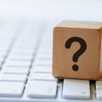Small wooden dice with black question mark symbol, viewed in close-up, sitting on top of white laptop computer keyboard, with blurred copy space